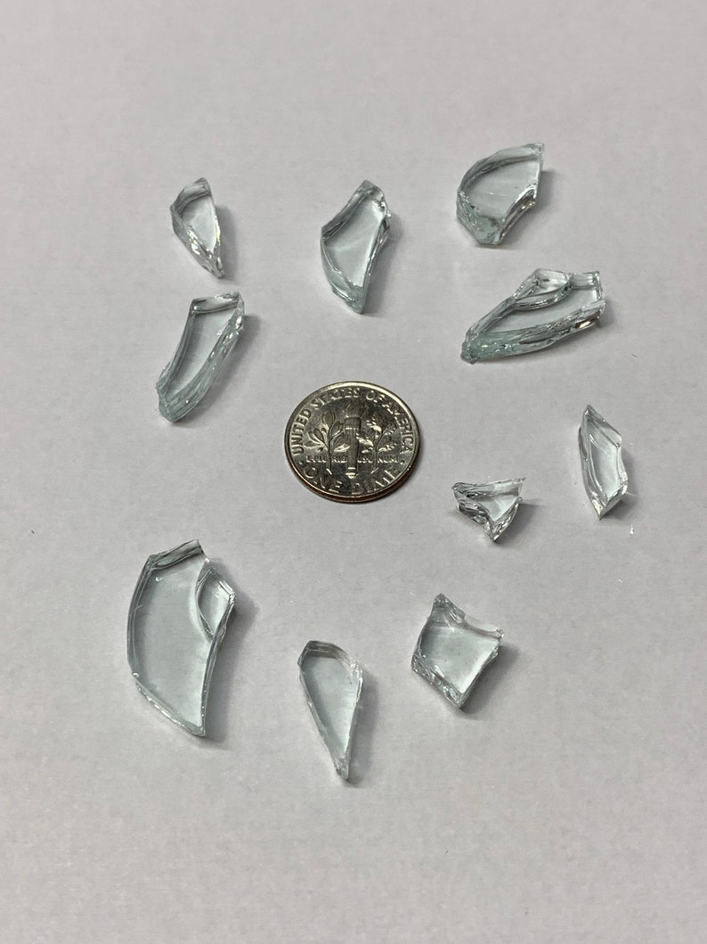 May include: A collection of clear, irregularly shaped glass shards scattered on a white surface. A U.S. dime is in the center of the image, providing a size reference for the fragments.