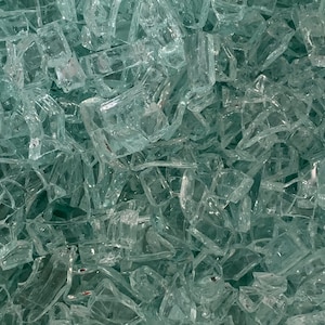 May include: Close-up of a pile of clear, irregularly shaped glass shards. The shards are translucent and reflect light, creating a sparkling effect. The pieces vary in size and shape, with sharp edges.