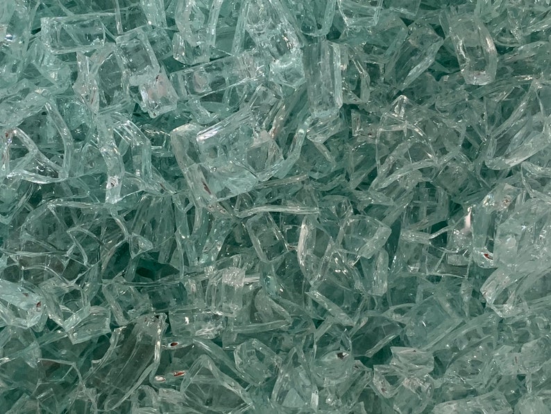 May include: Close-up of a pile of clear, irregularly shaped glass shards. The shards are translucent and reflect light, creating a sparkling effect. The pieces vary in size and shape, with sharp edges.
