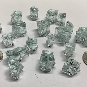 May include: A collection of clear, iridescent glass shards, some with a slight green tint, scattered on a white surface. The shards are various sizes and shapes, with some appearing more cube-like than others. Two US coins are included for scale.
