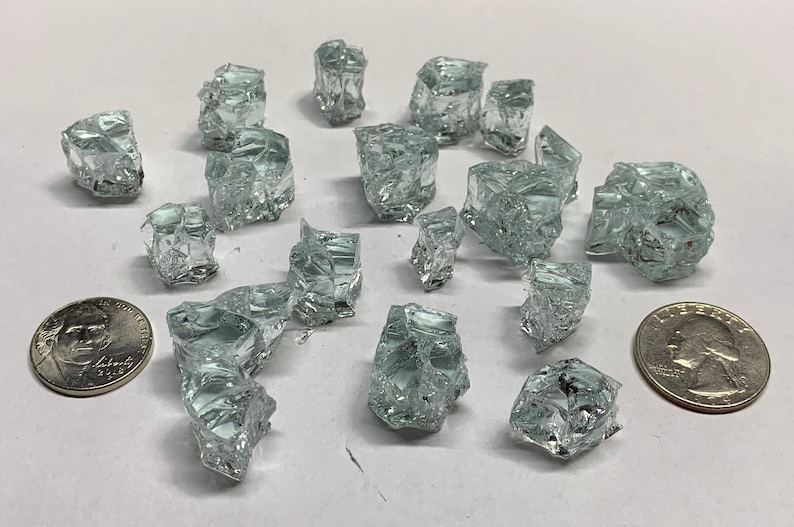 May include: A collection of clear, iridescent glass shards, some with a slight green tint, scattered on a white surface. The shards are various sizes and shapes, with some appearing more cube-like than others. Two US coins are included for scale.