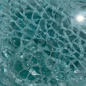 May include: A close-up of a cracked glass surface with a web-like pattern of fractures. The glass is a light blue-green color and reflects light.