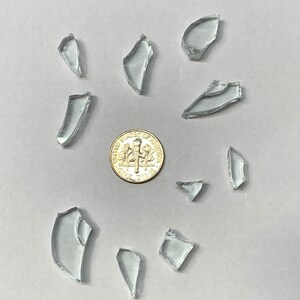 May include: A collection of clear, irregularly shaped glass shards scattered around a dime on a light gray surface. The shards vary in size and shape, reflecting light.