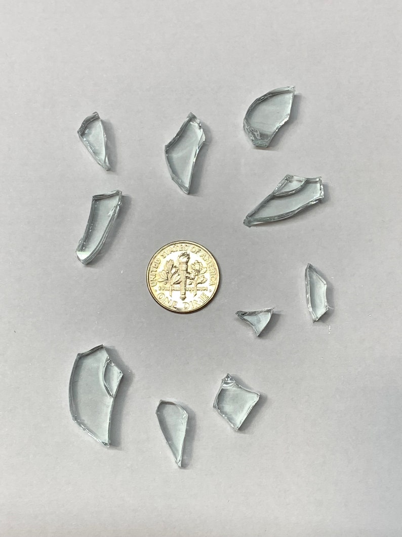 May include: A collection of clear, irregularly shaped glass shards scattered around a dime on a light gray surface. The shards vary in size and shape, reflecting light.