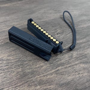 May include: A black plastic ammunition holder for .22 LR cartridges, featuring a removable cap with the text .22 LR embossed on the side. The holder contains a row of brass-colored cartridges and includes a black wrist lanyard attached to one end.