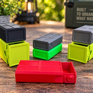 May include: A collection of rectangular storage containers with sliding drawers in black, lime green, dark gray, and red. Each container features a modular design with a grid pattern on the top surface and a circular side detail, arranged in a group.