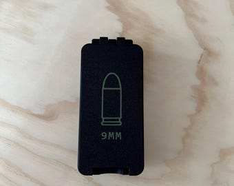 May include: A black rectangular object with a 9MM bullet graphic in light green. The object is on a light brown wooden surface.