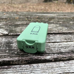 May include: A light green rectangular case with the text "9MM" printed on the front and top. The top of the case features a graphic of a bullet. The case is sitting on a weathered wooden surface.