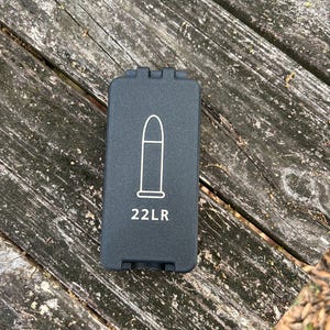 May include: A black rectangular object with a white outline of a bullet and the text "22LR". The object has a hinged top and is resting on a weathered wooden surface.