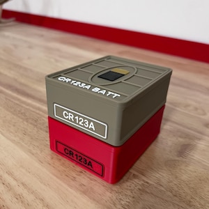 May include: A stack of two rectangular storage containers for CR123A batteries. The top container is tan with the text CR123A BATT and CR123A printed on it. The bottom container is red with the text CR123A printed on a black label.