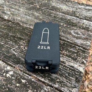 May include: A rectangular black plastic storage case for ammunition. The top lid features a white line drawing of a bullet and the text 22LR. The front latch also displays the text 22LR in white lettering against a black background.