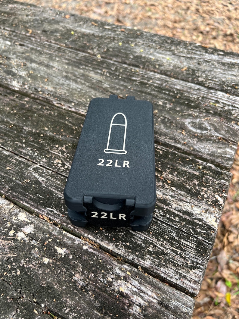 May include: A rectangular black plastic storage case for ammunition. The top lid features a white line drawing of a bullet and the text 22LR. The front latch also displays the text 22LR in white lettering against a black background.