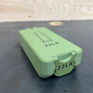 May include: A green rectangular container with a hinged lid. The lid features a black outline of a bullet and the text "22LR". The same text is also on the front of the container.