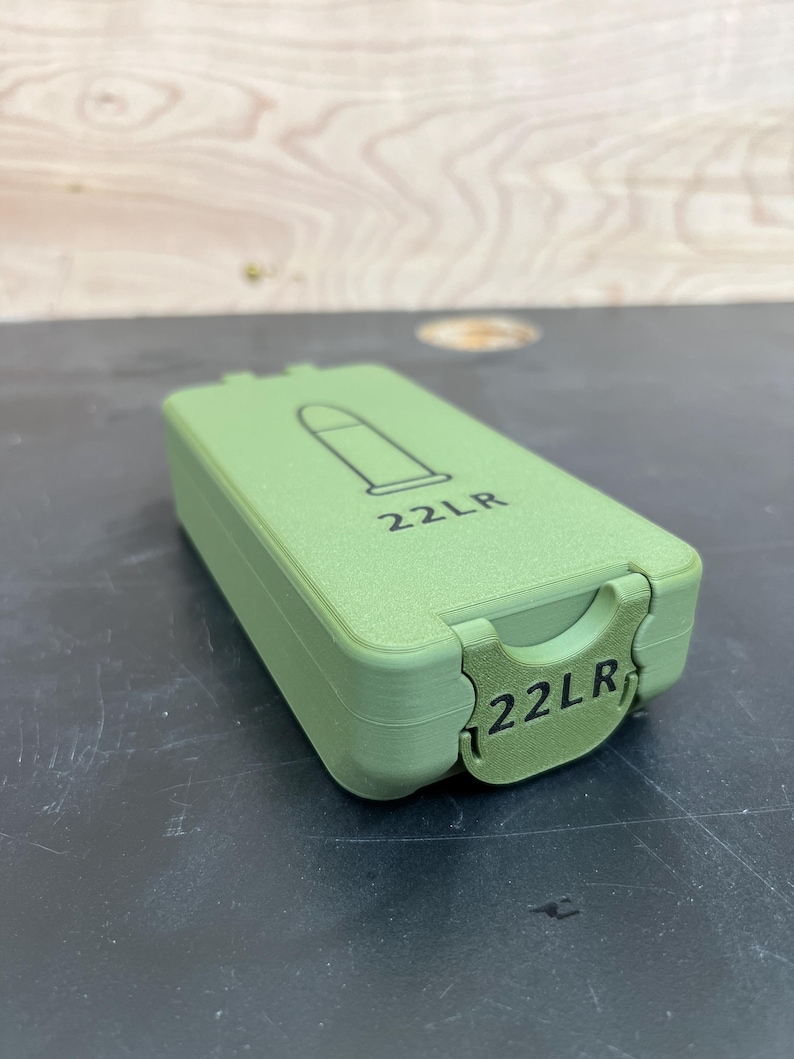 May include: A green rectangular container with a hinged lid. The lid features a black outline of a bullet and the text "22LR". The same text is also on the front of the container.