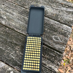 May include: An open, black rectangular case with a grid of small, gold-colored objects inside. The case is on a weathered wooden surface, suggesting outdoor use. The objects appear to be small, round, and metallic.
