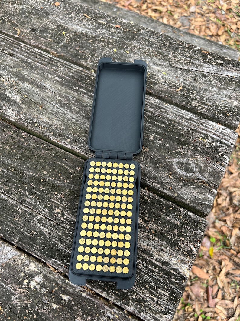 May include: An open, black rectangular case with a grid of small, gold-colored objects inside. The case is on a weathered wooden surface, suggesting outdoor use. The objects appear to be small, round, and metallic.