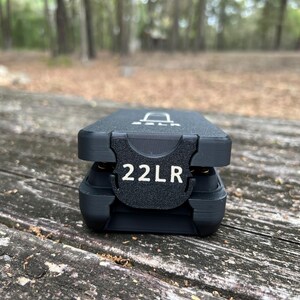 May include: A black 22LR ammunition case with the text "22LR" in white. The case is rectangular with a textured surface and a hinged lid. The case is sitting on a weathered wooden surface.