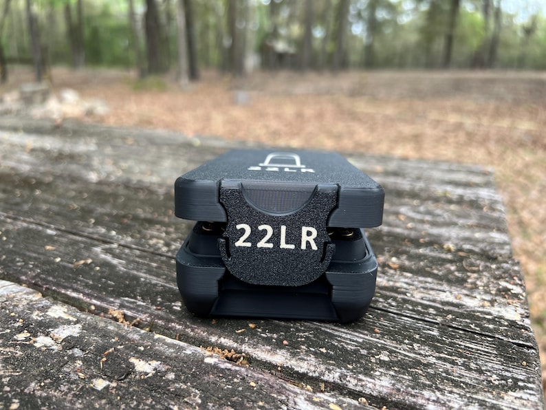 May include: A black 22LR ammunition case with the text "22LR" in white. The case is rectangular with a textured surface and a hinged lid. The case is sitting on a weathered wooden surface.