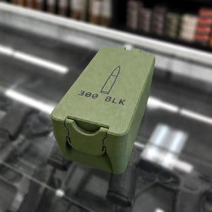 May include: An olive green rectangular container with a hinged lid. The lid features a simple line drawing of a bullet and the text "300 BLK" in black. The container has a clasp closure and appears to be made of a durable material.