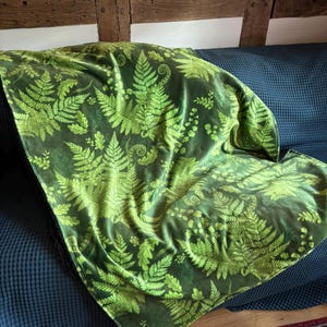 May include: A green fern-patterned blanket on a blue sofa. The blanket displays a repeating design of fern fronds in different green shades. The sofa is against a white wall with wooden beams.