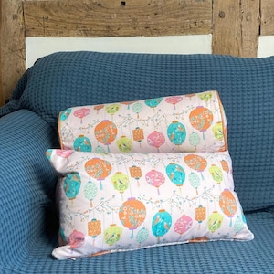 May include: Two decorative cushions with a white background and a pattern of colourful lanterns and flowers. The cushions are on a blue sofa.