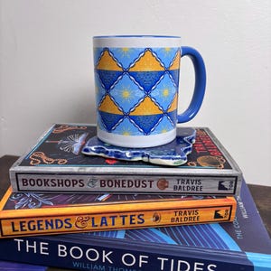 May include: A blue and white ceramic mug with a blue handle, featuring a repeating pattern of yellow suns and blue water designs. The mug sits on a matching coaster, atop a stack of books with titles like "Bookshops & Bonedust" and "Legends & Lattes."