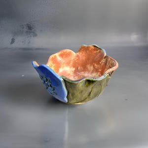 Könnte beinhalten: Eine handgefertigte Keramikschüssel mit einem wellenförmigen, blumenförmigen Rand. Die Schüssel ist in Blautönen, Grün- und Orangetönen glasiert.