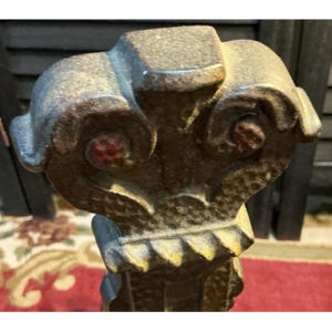 May include: Close-up of a decorative, ornate metal object with a weathered, aged appearance. The object features intricate scrollwork and a square top, with hints of red and gold accents. The overall design suggests a vintage or antique aesthetic.