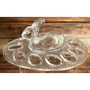 Puede incluir: Una fuente de vidrio transparente para servir huevos de Pascua con un centro de mesa en forma de conejo. La fuente tiene una superficie texturizada y hendiduras en forma de huevo alrededor del borde. El conejo tiene detalles, incluyendo orejas y cuerpo.