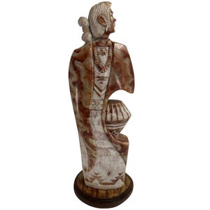 May include: A carved stone sculpture of a Native American figure, featuring intricate details and a natural color palette. The figure is adorned with a necklace and traditional clothing, holding a pot. The sculpture is mounted on a wooden base.