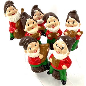 May include: A collection of seven garden gnomes, each with a white beard, brown hat, and red jacket. Some gnomes are playing musical instruments, including a guitar, accordion, and double bass. The gnomes have green trousers and are standing on a white surface.