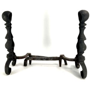 May include: A pair of black metal fireplace andirons with ornate details. The andirons are connected by a metal bar.
