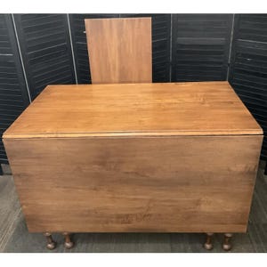 May include: A light brown wooden drop-leaf table with a matching rectangular leaf propped up behind it. The table has a rectangular top and rounded legs. The table is made of wood and has a natural finish.