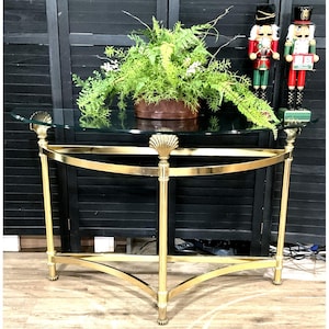 May include: A gold-toned console table with a glass top and seashell accents. The table features curved legs and a lower shelf. A potted plant and two decorative nutcrackers are on the table.