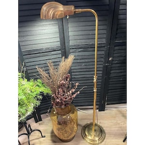 May include: A tall brass floor lamp with a curved, shell-shaped shade and a slender, adjustable neck. The lamp features a circular base and is positioned next to a large, amber-colored glass vase filled with dried brown botanical stems.
