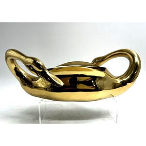 May include: A gold-colored swan-shaped decorative bowl. The bowl features two swan heads and necks forming handles. The polished surface reflects light, creating a shiny appearance. The bowl is displayed on a clear acrylic stand.