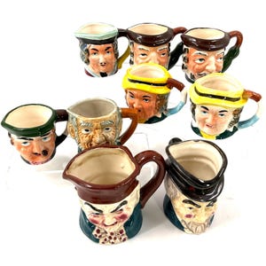 Puede incluir: Colección de tazas de cerámica vintage, cada una con forma y pintada de forma única para parecerse a una cara humana. Las tazas presentan diversas expresiones faciales y están adornadas con sombreros y detalles coloridos. Las asas son marrones, sobre fondo blanco.