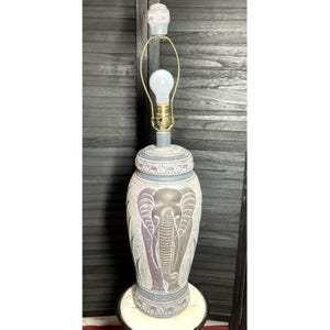 May include: A decorative table lamp with an elephant design. The lamp features a large, urn-shaped base with an etched elephant motif in shades of purple and gray. The lamp has a light bulb and a gold-colored metal frame. The lamp is approximately 24 inches tall.