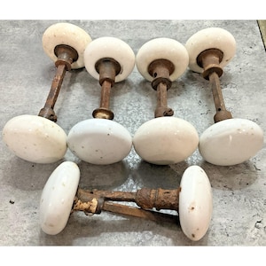 May include: A collection of five vintage door knobs featuring smooth, round white porcelain handles attached to weathered, rusted metal spindles and hardware components. The set includes four upright standing knobs and one horizontal knob.