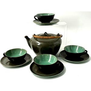 Puede incluir: Un juego de té de cerámica con una tetera y cuatro tazas y platillos. La tetera es de color verde oscuro con un asa tejida. Las tazas y platillos son de color verde oscuro con un interior turquesa.