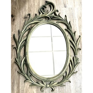 May include: Oval-shaped wall mirror with an ornate, olive-green frame. The frame features a wheat-like design around the mirror's edge and decorative flourishes at the top and bottom. The mirror is set against a light-colored wooden surface.