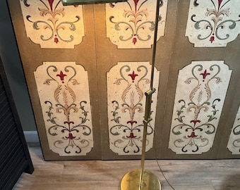 May include: A brass floor lamp with a cylindrical head and a slender, adjustable vertical stand. The lamp features a circular base and a metallic finish, emitting a warm light from the horizontal shade positioned at the top of the frame.