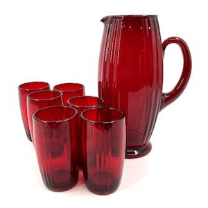 Puede incluir: Un juego de seis vasos rojos y una jarra a juego. La cristalería es de color rojo intenso y tiene detalles acanalados verticales. La jarra tiene un asa grande y un pico vertedor. El juego es probablemente para servir bebidas.