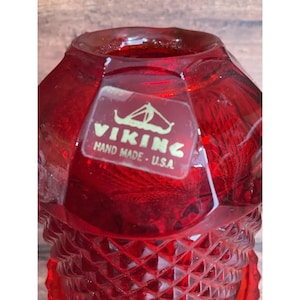 Pode incluir: Close-up de um objeto de vidro vermelho rubi com um padrão de diamante na parte inferior. O objeto tem um adesivo que diz "VIKING HAND MADE - U.S.A." com um logotipo de veleiro. O vidro é facetado e reflete a luz.