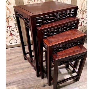 May include: A set of four dark brown wooden nesting tables with rectangular tops and straight legs. Each table features an openwork apron with a central circular medallion design. The tables are stacked in descending order of size.