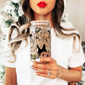 May include: A clear glass tumbler with a bamboo lid and straw. The tumbler features a black line drawing of a mother and child embracing, with floral and heart accents. The design is set against a frosted background.