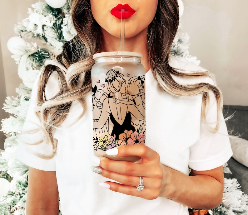 May include: A clear glass tumbler with a bamboo lid and straw. The tumbler features a black line drawing of a mother and child embracing, with floral and heart accents. The design is set against a frosted background.