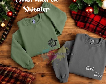 Könnte beinhalten: Zwei bestickte Pullover in Olivgrün und Grau werden auf einer Holzoberfläche präsentiert. Der olivgrüne Pullover hat ein schwarzes Sonnendesign, während der graue Pullover die Initialen "S.W. D.W." in Weiß hat. Der Text "Embroidered Sweater" steht oben.