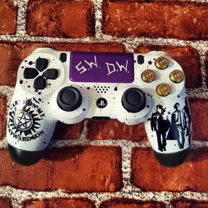 Può includere: Un controller PlayStation 4 bianco con una placca frontale viola e pulsanti neri. Il controller ha un design in bianco e nero con un simbolo in bianco e nero sul lato sinistro e una silhouette in bianco e nero di quattro persone sul lato destro. La placca frontale ha il testo "S.W. D.W." scritto in bianco. Il controller ha bossoli di proiettile dorati sulla parte superiore.