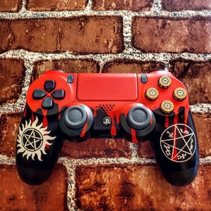 Puede incluir: Un mando de PlayStation 4 rojo y negro con detalles dorados y un símbolo de Supernatural a cada lado. El mando tiene un diseño de pintura roja que gotea.